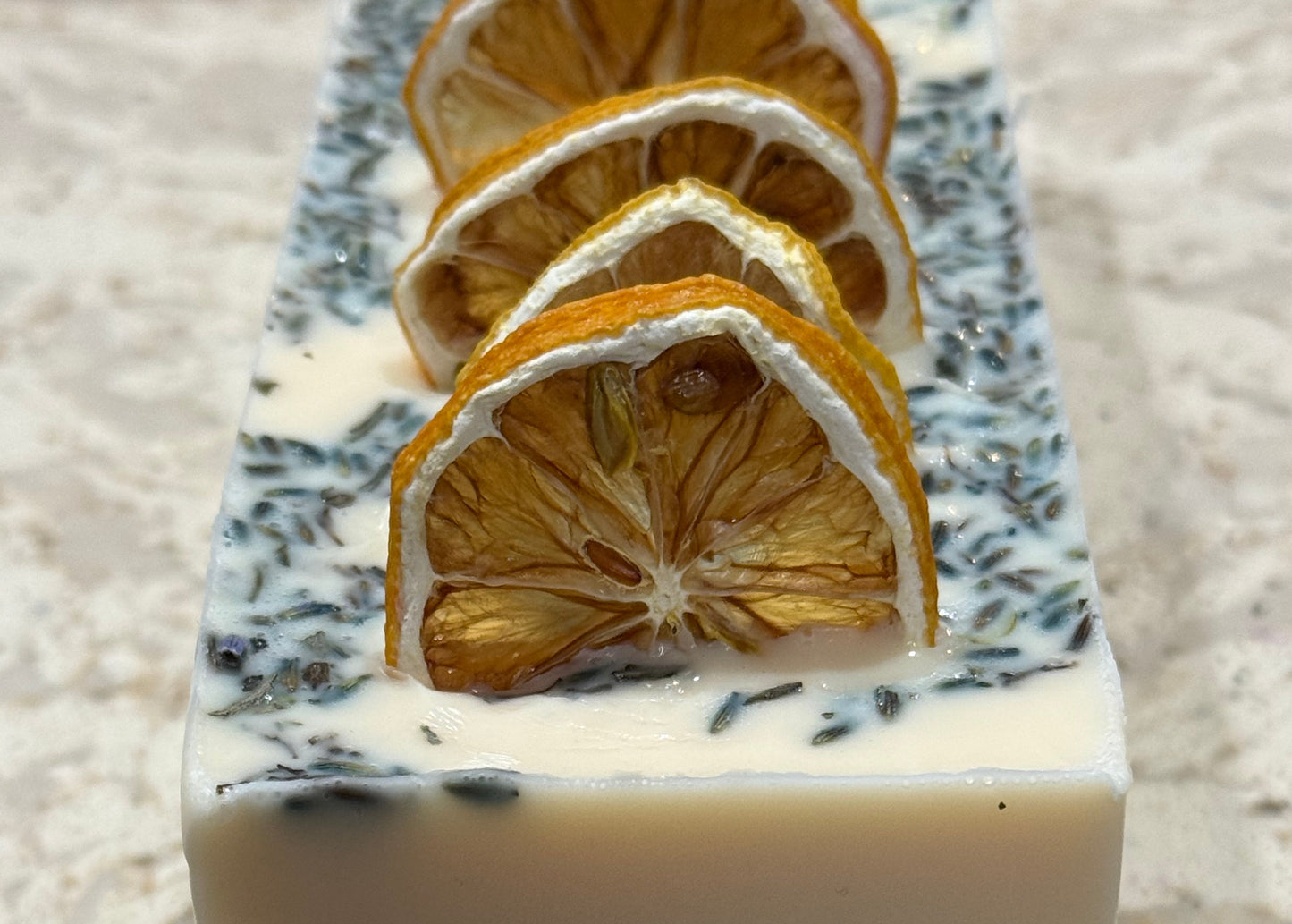 Square block of soap with dried orange slices embedded, on a textured surface.