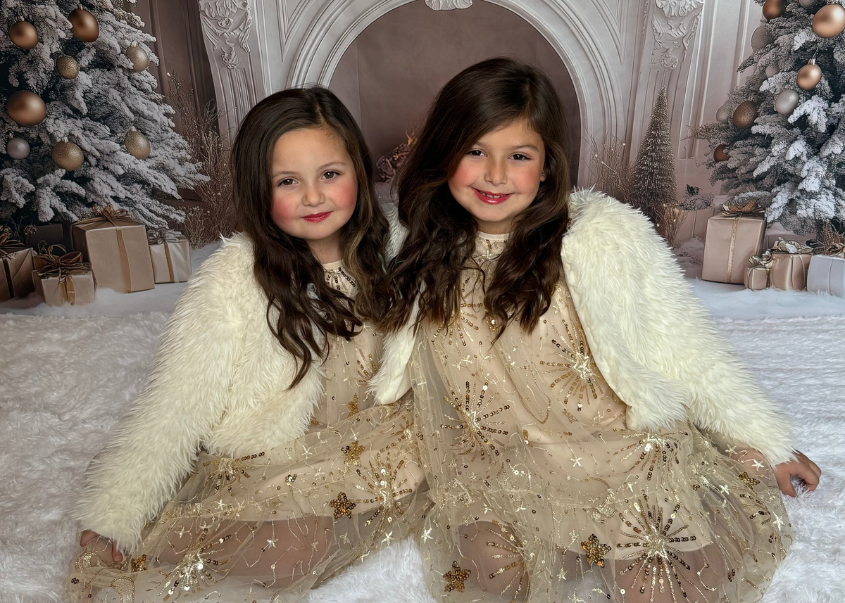 Two young girls in festive outfits sitting in front of a decorated fireplace with Christmas trees.